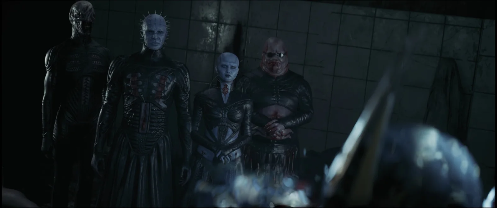 Four Cenobites standing shoulder to shoulder in a dark tiled room, watching, with the puzzle box blurred in the foreground