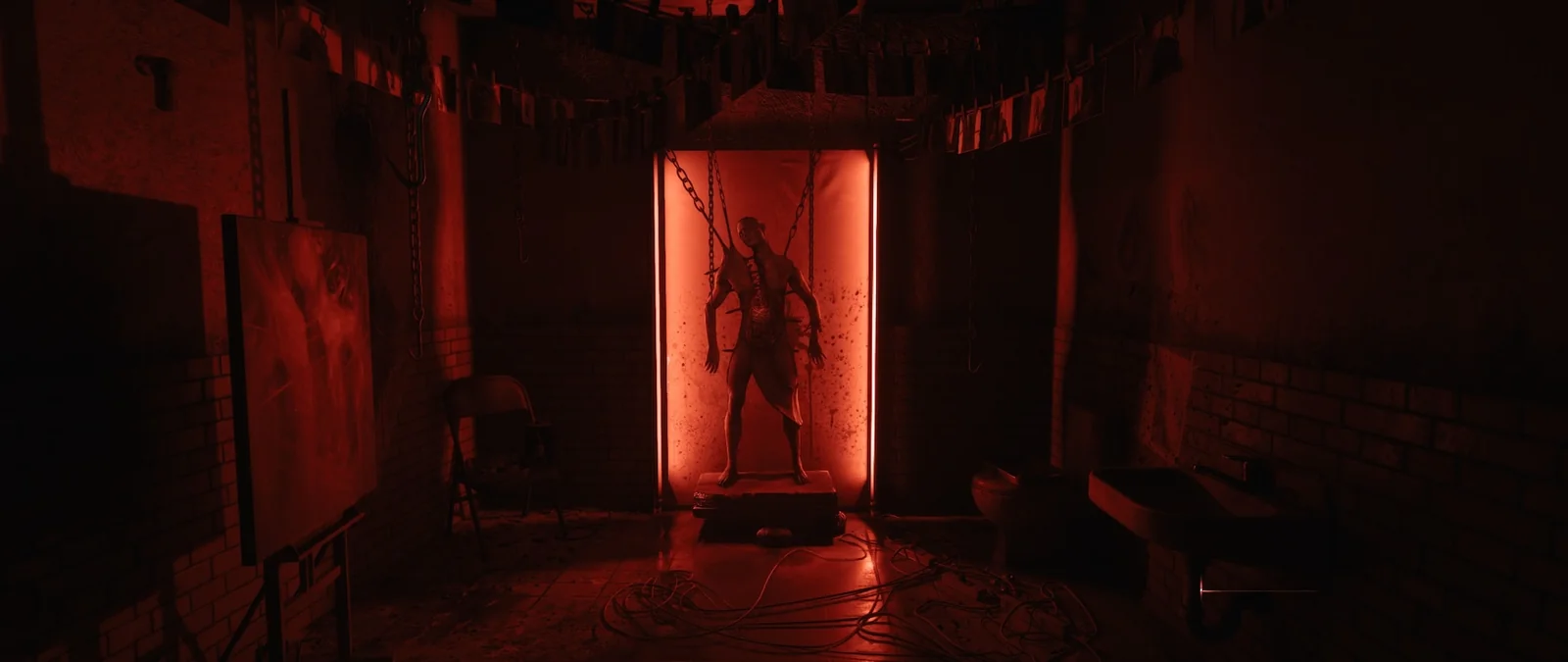 A flayed figure hung on chains inside a red-lit glass booth, in a grimy studio with a canvas on an easel