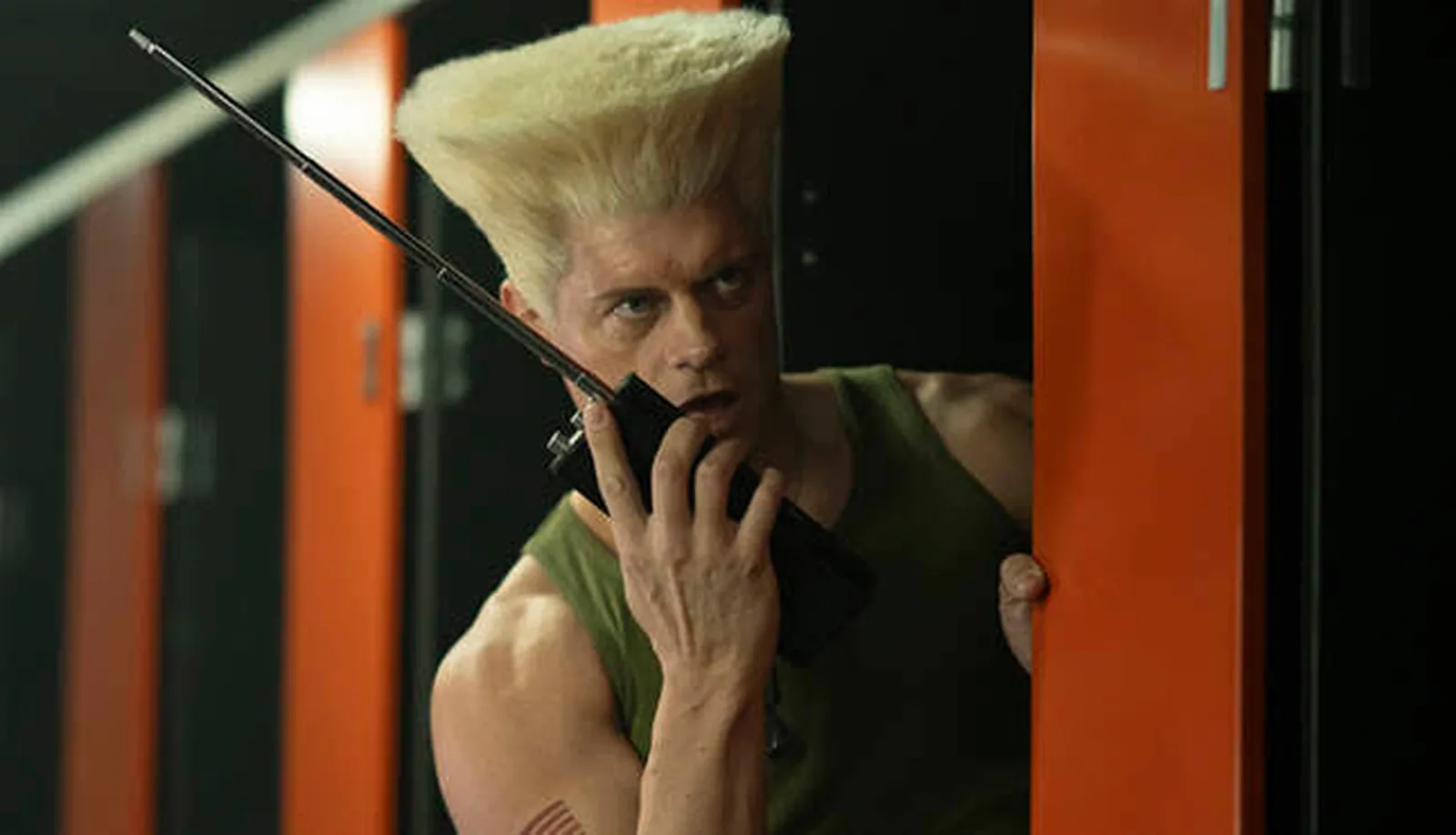 Cody Rhodes as Guile, flat-top towering, talking into a walkie-talkie in a green tank top