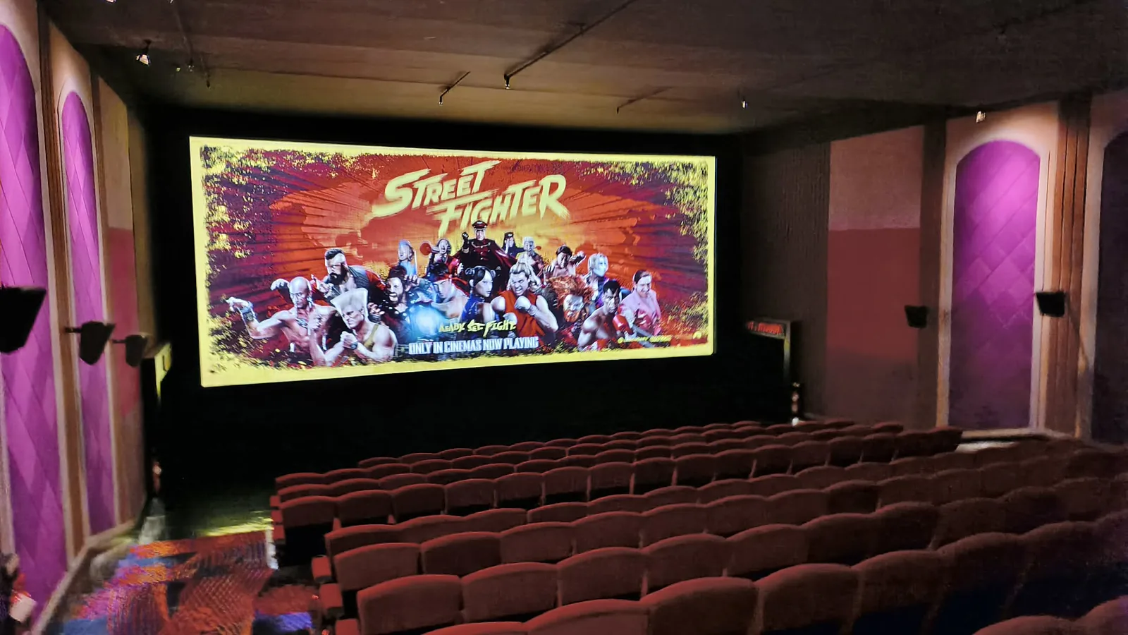 A movie theater’s screen lit up with the Street Fighter poster, the whole roster crowded under the logo above rows of red seats