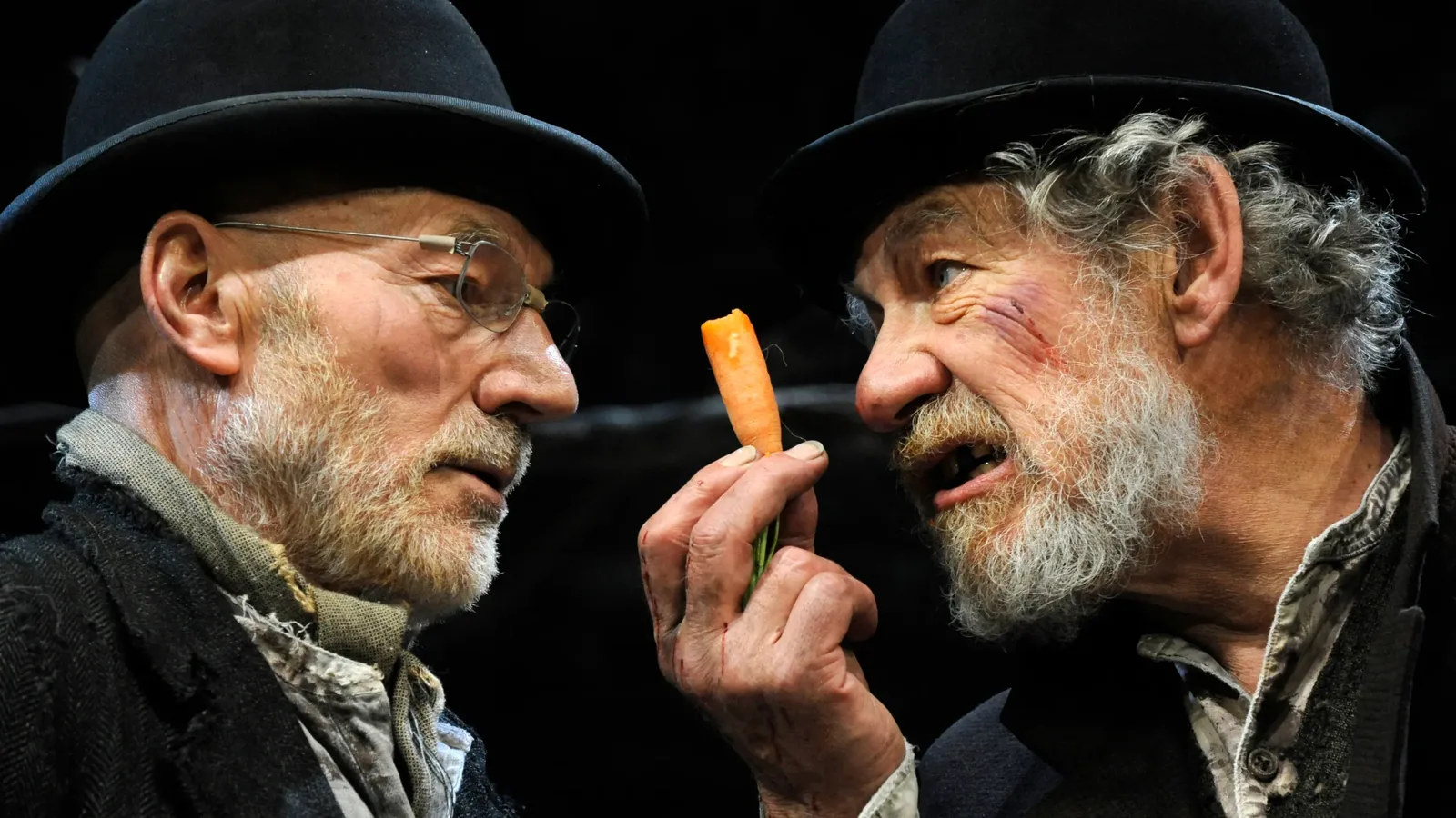 Patrick Stewart and Ian McKellen in bowler hats and ragged coats in Waiting for Godot, McKellen holding up a carrot between them