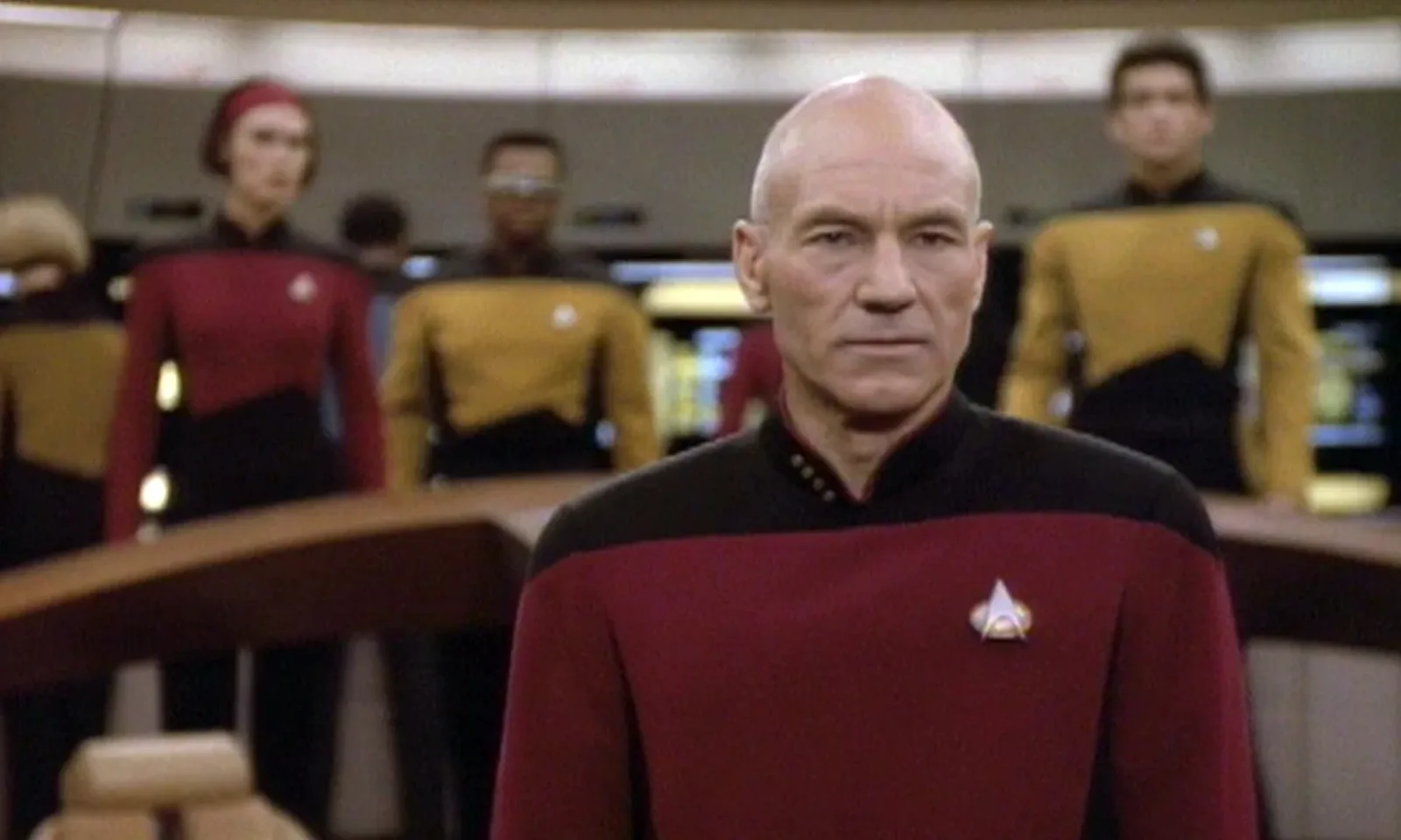 Patrick Stewart as Captain Jean-Luc Picard in his red uniform on the bridge of the Enterprise, his crew standing behind him