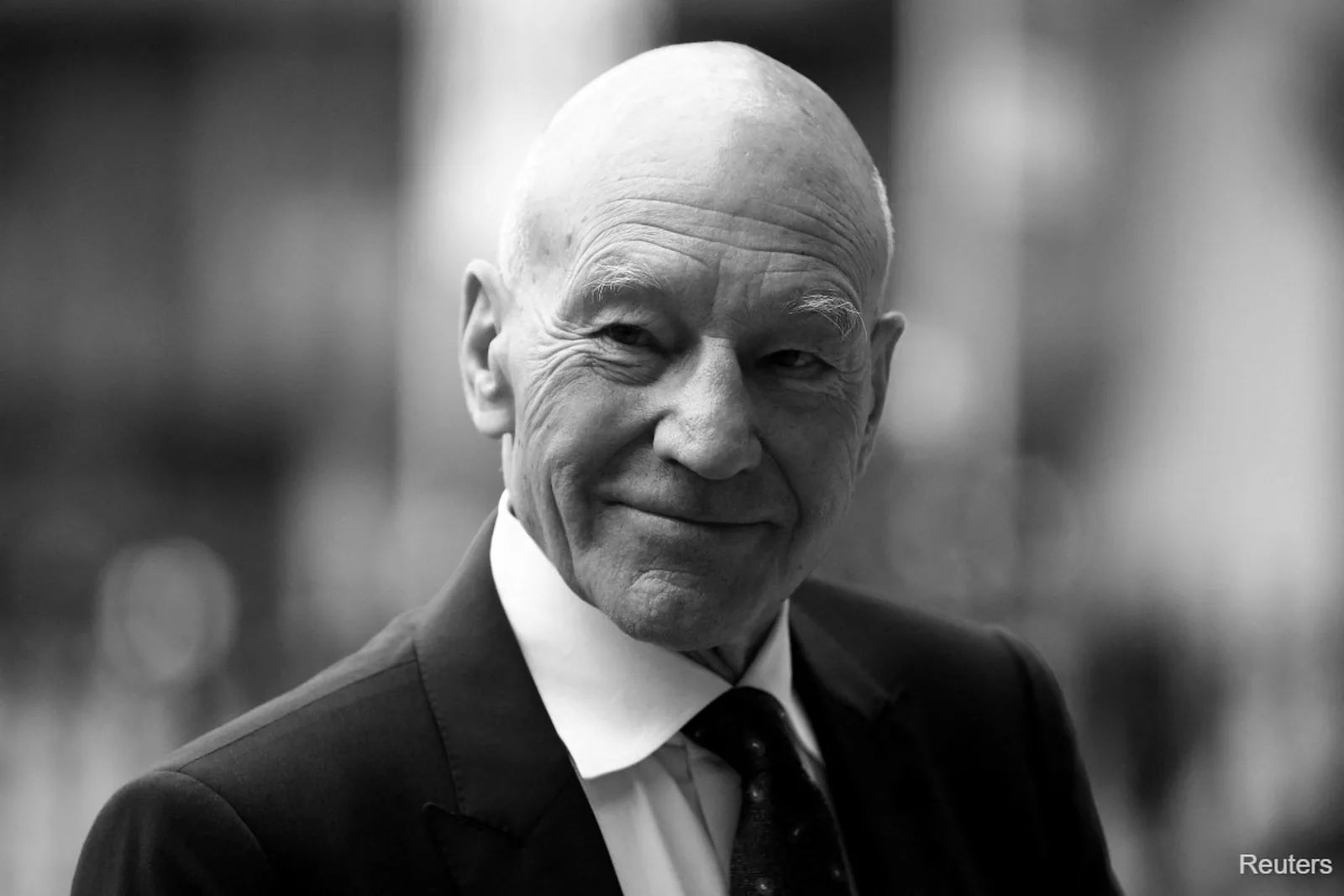 A black-and-white portrait of Sir Patrick Stewart in a dark suit and tie, smiling warmly