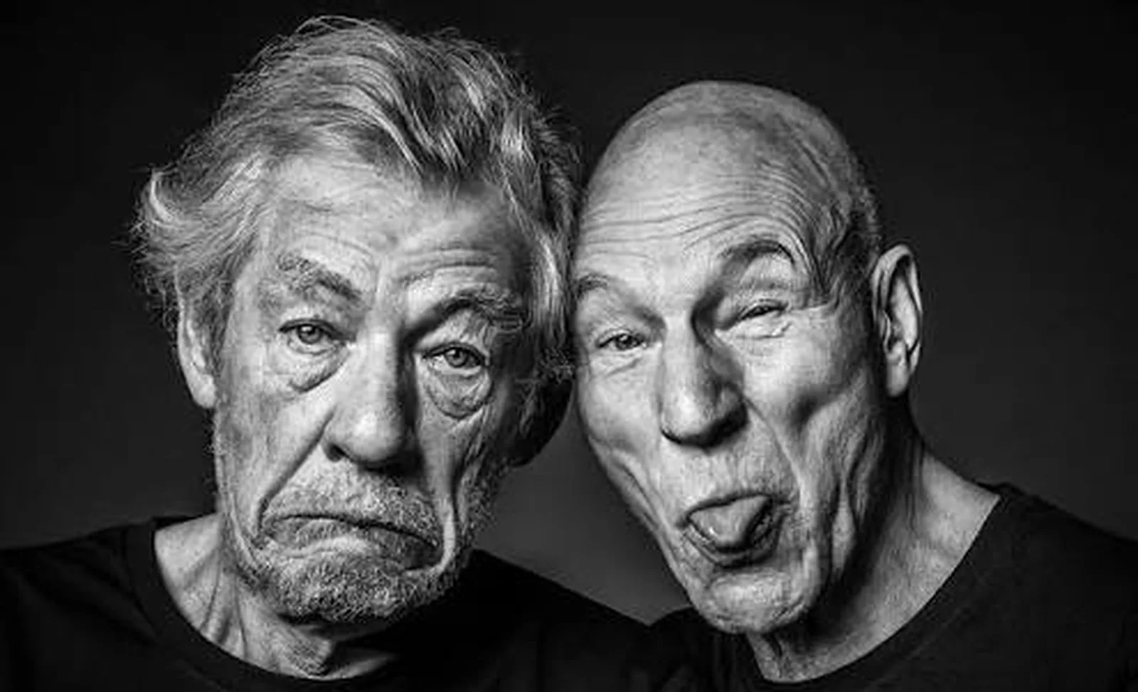 A black-and-white portrait of Patrick Stewart and Ian McKellen with their heads together, McKellen scowling and Stewart sticking out his tongue