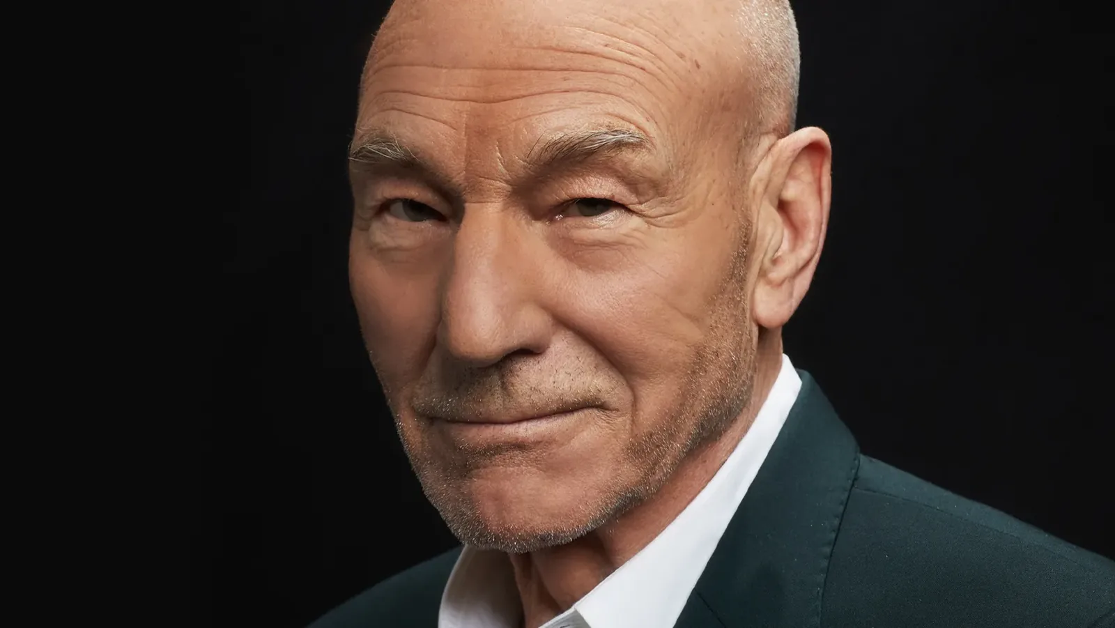 Sir Patrick Stewart in a dark suit against a black background, smiling slightly